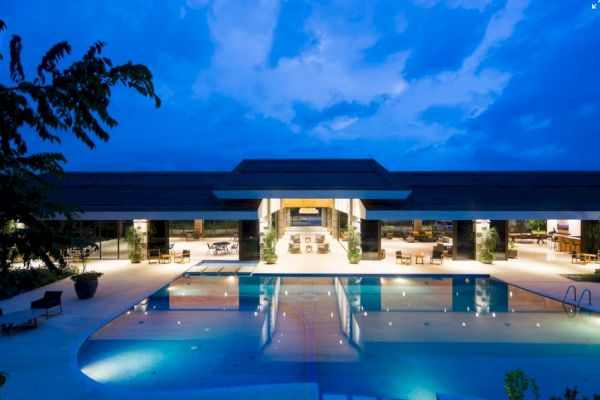 A luxurious building with a wide open area, featuring a large illuminated swimming pool under a blue evening sky.