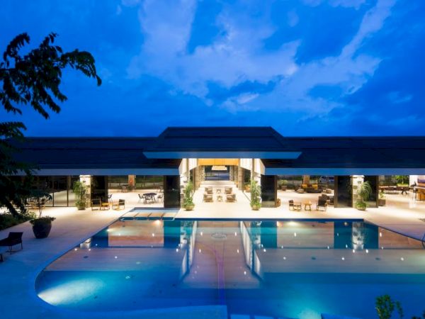 A luxurious building with a wide open area, featuring a large illuminated swimming pool under a blue evening sky.