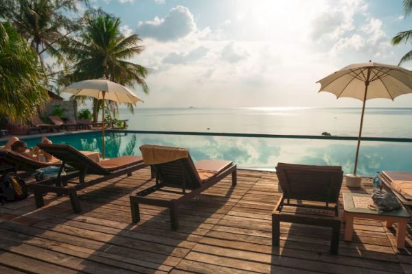 A serene poolside scene with lounge chairs, umbrellas, and palm trees overlooking a tranquil ocean view under a partly cloudy sky.