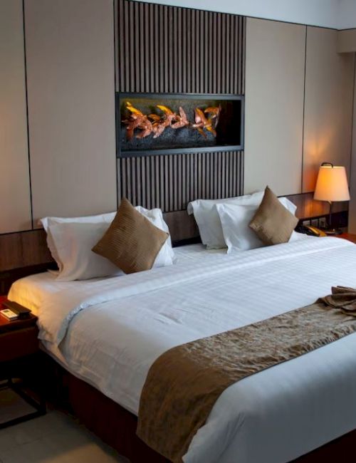 A cozy hotel room with a large bed, two bedside lamps, decorative cushions, and a folded towel on the bed with a wall art piece above.