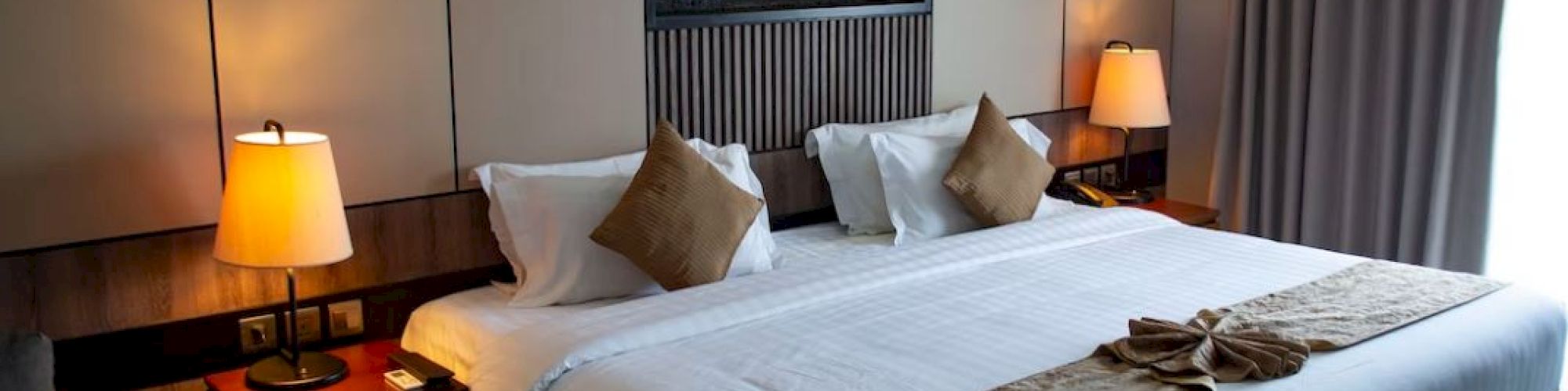 The image shows a hotel room with a neatly made bed, bedside tables with lamps, and a modern design with neutral tones and accent pillows.