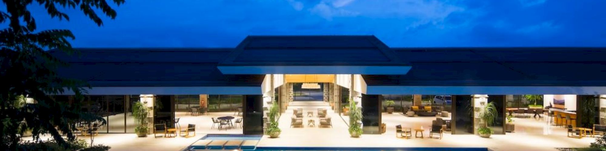 Luxurious building with large illuminated pool at night, surrounded by seating areas and trees, and a vibrant blue sky above.