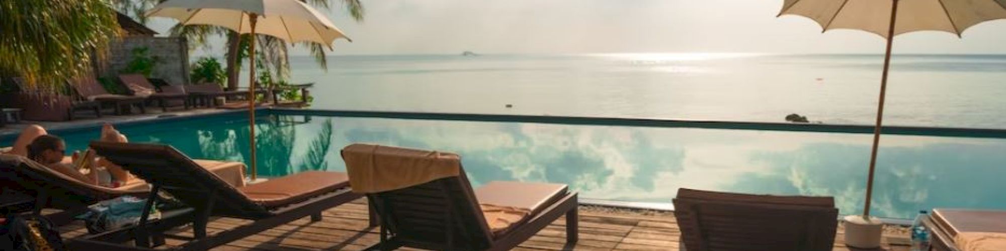 A serene poolside view with sun loungers, umbrellas, palm trees, and a tranquil ocean under a partly cloudy sky.