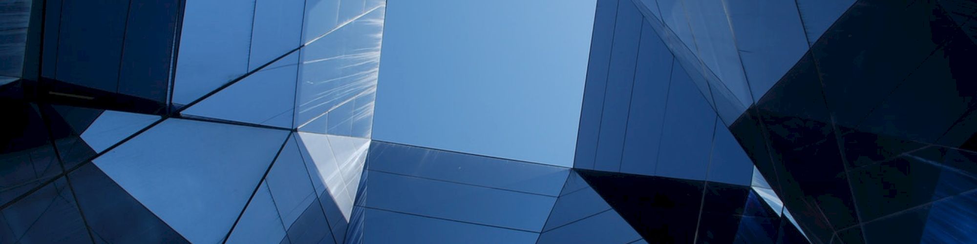 A view looking up at a geometric structure with reflective glass surfaces and a blue sky in the center.