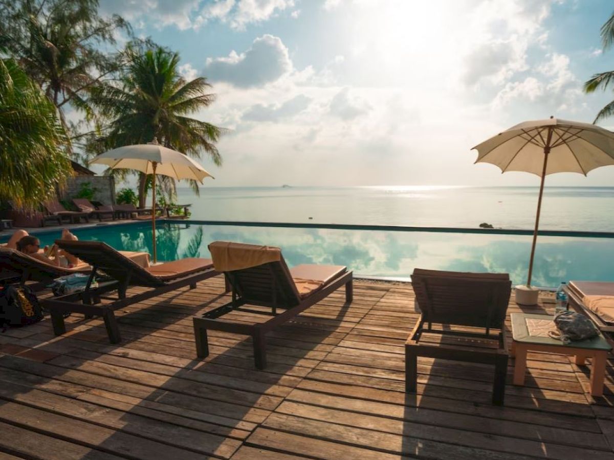 A serene poolside scene with lounge chairs, umbrellas, and palm trees overlooking a calm, sunny ocean view, creating a relaxing atmosphere.