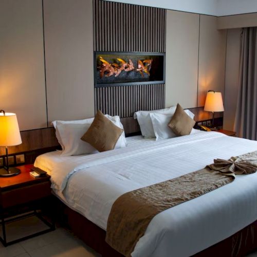 A neatly arranged hotel room with a king-sized bed, warm lighting, cushions, a decorative towel, and art on the wall.