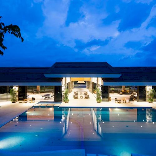 A luxurious building with a large pool at night, surrounded by lounge chairs, under a blue, partly cloudy sky.