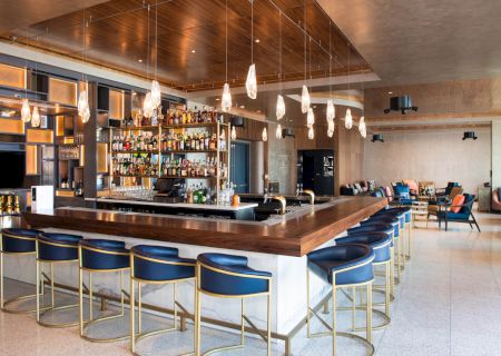 A modern bar with a wooden counter, blue cushioned stools, hanging lights, and a well-stocked liquor shelf in a spacious lounge area.