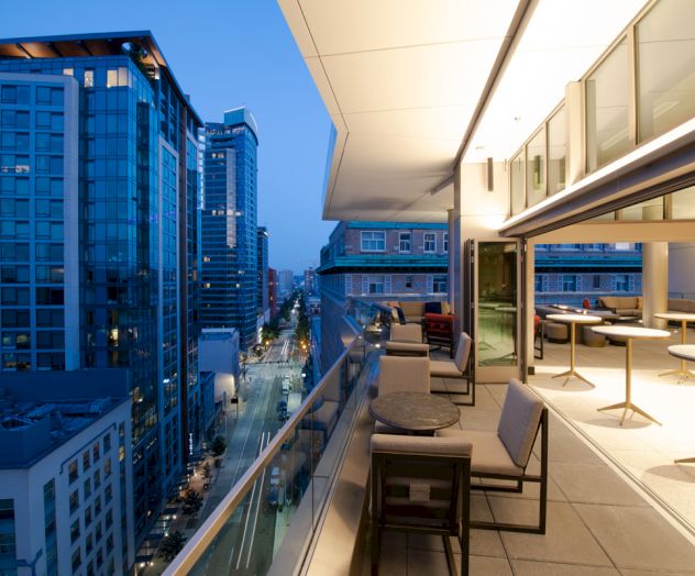 A modern urban balcony overlooking a city street, with seating areas, tall buildings, and illuminated interior space at dusk.