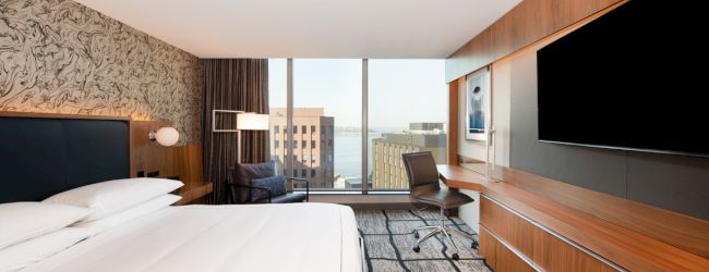 A modern hotel room with a large bed, desk with chair, and flat-screen TV. It features floor-to-ceiling windows and a city view.