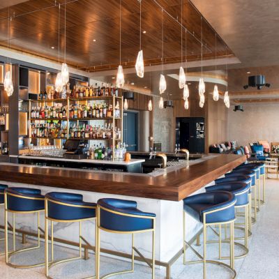 This image shows a modern bar with a wooden countertop, blue stools, hanging lights, and a well-stocked display of bottles in a spacious setting.