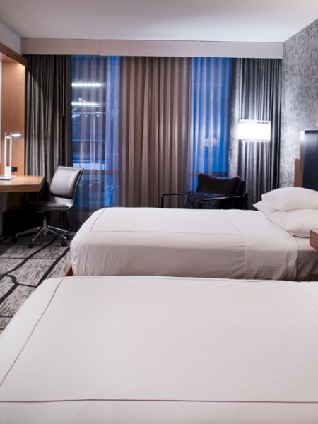 The image shows a modern hotel room with two beds, a flat-screen TV, a desk, and stylish décor, lit by lamps on a patterned wall.