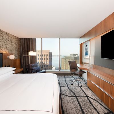 A modern hotel room with a large bed, wall-mounted TV, desk, and a chair by the window offering a city view.
