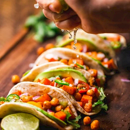 The image shows tacos filled with chickpeas, avocado, and vegetables, being topped with a squeeze of lime on a wooden board.