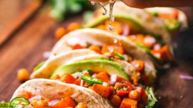The image shows tacos filled with chickpeas, avocado, and vegetables, being topped with a squeeze of lime on a wooden board.