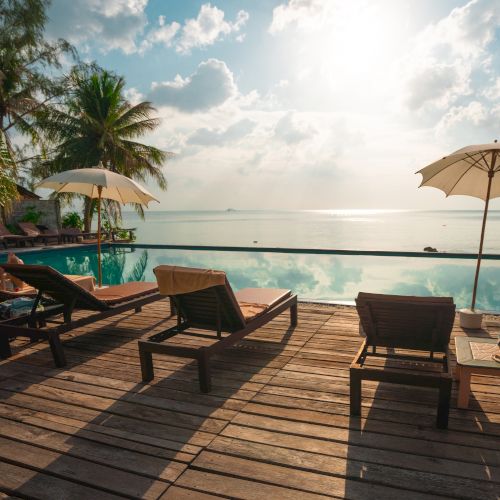A serene beachfront scene with lounge chairs, umbrellas, and palm trees by a pool, overlooking calm ocean waters under a bright sky.
