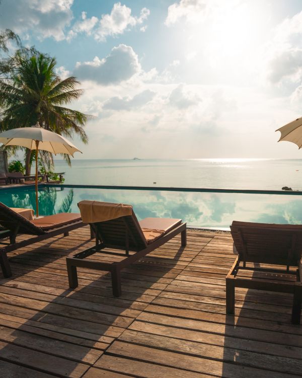 A serene beachfront scene with lounge chairs, umbrellas, and palm trees by a pool, overlooking calm ocean waters under a bright sky.