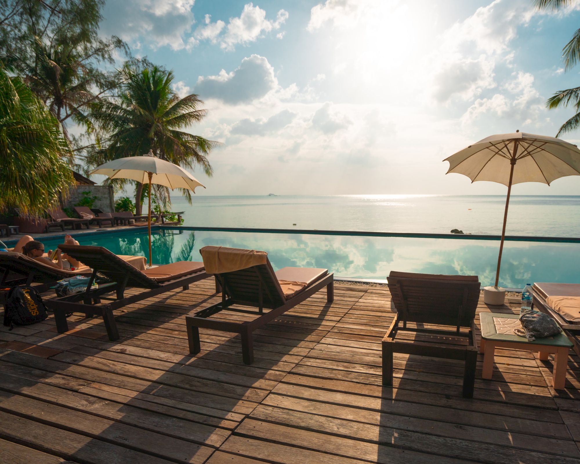 A serene poolside view with lounge chairs, umbrellas, and palm trees overlooking a calm sea under a partly cloudy sky.