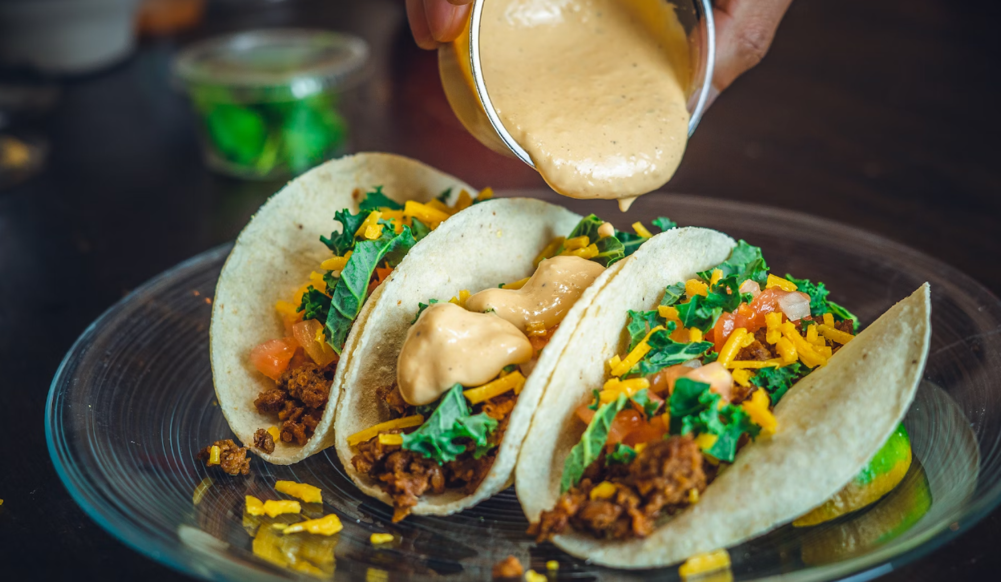 Three tacos with toppings like meat, cheese, tomatoes, and greens are on a plate, with sauce being poured over them.