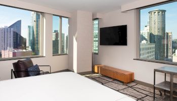 A hotel room with a bed, armchair, TV, and large windows showing a cityscape with tall buildings.