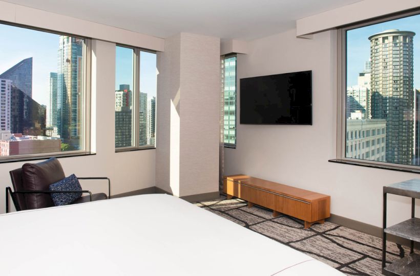 A hotel room with a bed, armchair, TV, and large windows showing a cityscape with tall buildings.