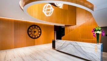 A modern lobby with a large clock, a marble reception desk, wood paneling, a stylish spiral staircase, and decorative lighting.