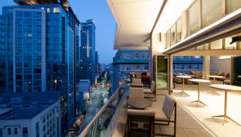 The image shows a modern cityscape with tall buildings and a spacious balcony featuring tables and chairs, overlooking a street at dusk.