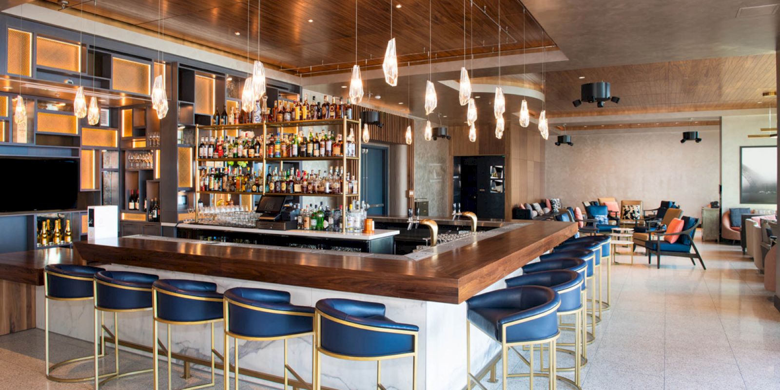 A modern bar with blue high chairs, a variety of bottles on shelves, and pendant lights, located in a spacious, stylish setting.
