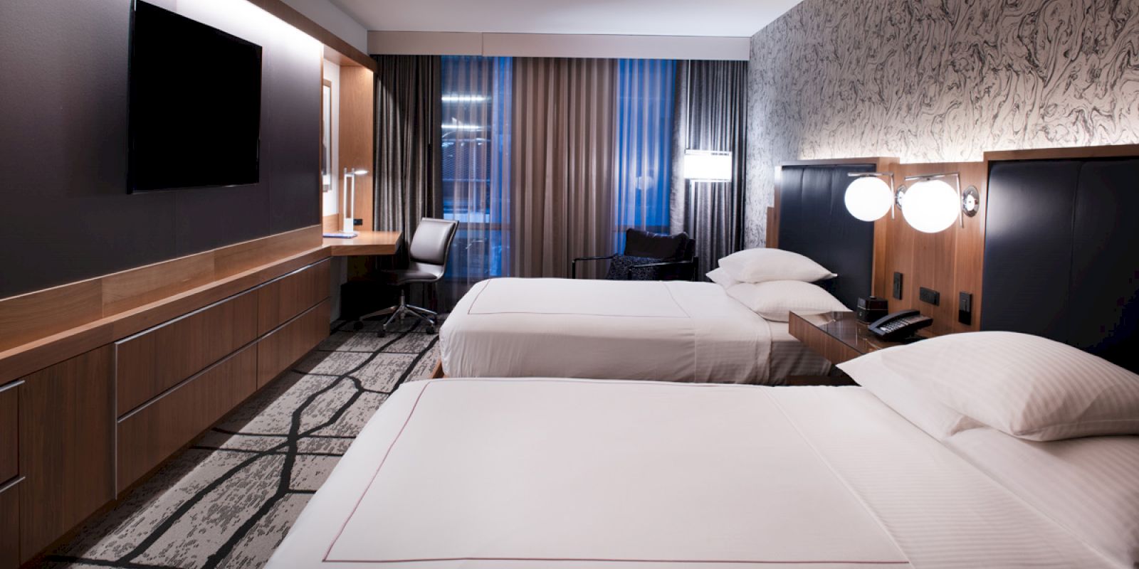 A modern hotel room with two beds, a desk, a large TV, and stylish lighting and decor, featuring patterned walls and curtains.