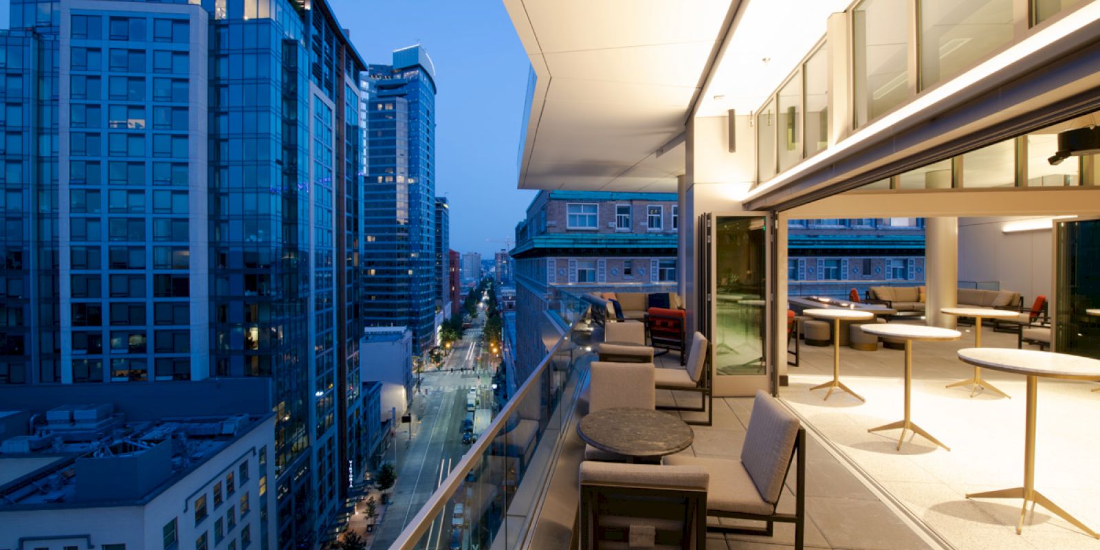 A modern urban patio with tables and chairs overlooks a cityscape of tall buildings at dusk, featuring a bright, open indoor space.