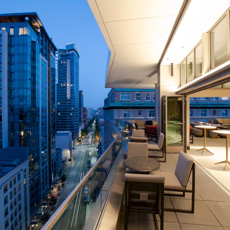 A modern cityscape featuring a sleek balcony with tables and chairs, overlooking illuminated skyscrapers and a bustling street below.