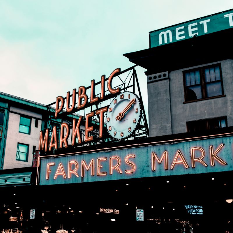 The image shows a public market sign with a clock and "Farmers Market" in neon. Buildings are in the background under a cloudy sky.