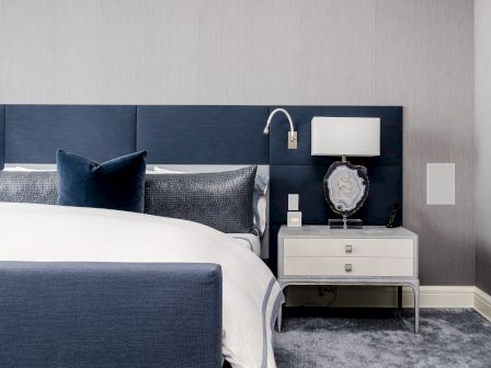 A modern bedroom with a navy blue bed, gray bedding, a nightstand, a lamp, decorative slice, and a textured wall background.