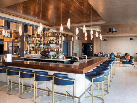 Modern bar with blue stools, wooden accents, and a fully stocked bar. Pendant lights hang from the ceiling. Cozy seating area nearby.