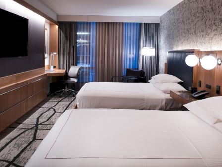 A modern hotel room with two double beds, a desk, chair, TV, and stylish lighting, featuring a patterned carpet and wall design.