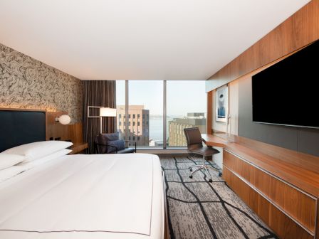 A modern hotel room with a large bed, TV, desk, chair, and a window offering a city view is in the image.