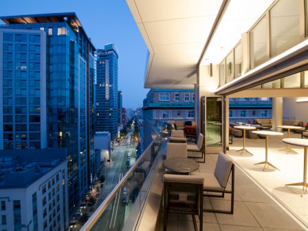 A modern cityscape at dusk with tall buildings and an elegant outdoor balcony featuring tables and chairs, overlooking a vibrant street.