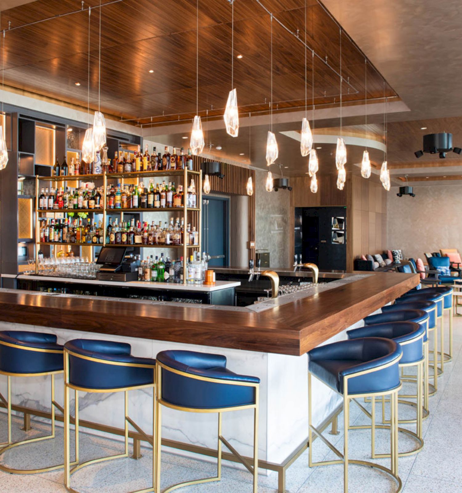 This image shows a modern bar with blue stools, pendant lights, and a well-stocked bar. There’s a cozy seating area in the background.
