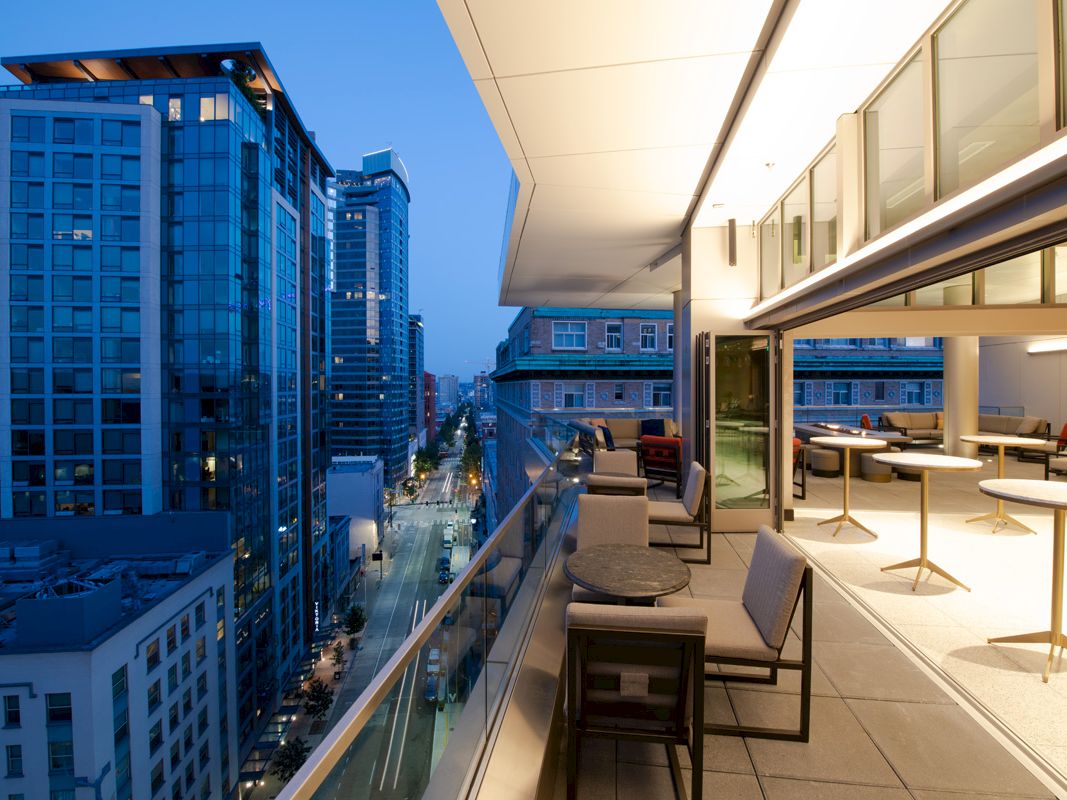 A modern rooftop terrace with tables and chairs overlooks a city skyline at dusk, featuring tall buildings and illuminated streets.