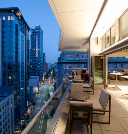 A modern rooftop patio with seating overlooks a city street and tall buildings at dusk, featuring sleek outdoor furniture and glass railings.