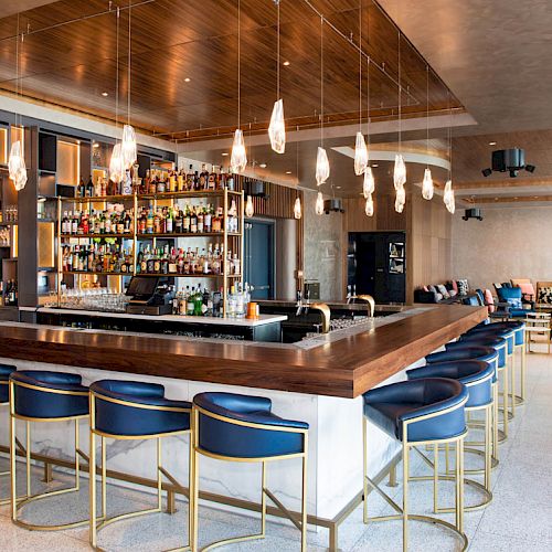 This image shows a modern bar with a wooden counter, surrounded by blue stools, and a sleek, well-stocked bar display with hanging lights.