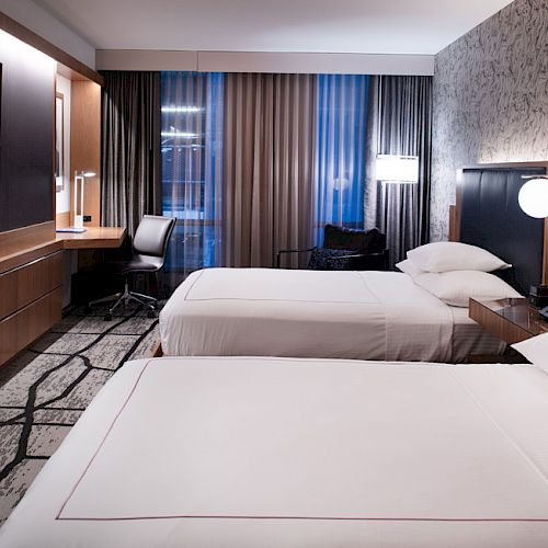 A modern hotel room with two beds, a wall-mounted TV, a desk, and a patterned carpet. The room has a sleek and contemporary design.