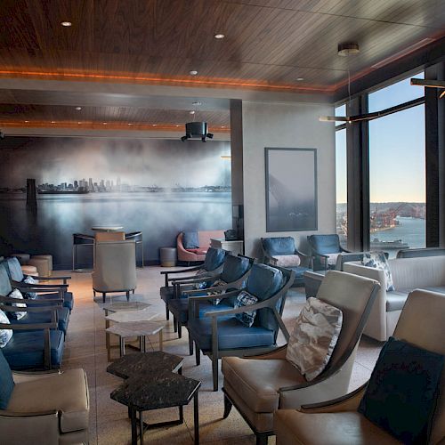 The image shows a modern lounge area with chairs, a large mural, and windows overlooking a cityscape and waterfront view, including a Ferris wheel.