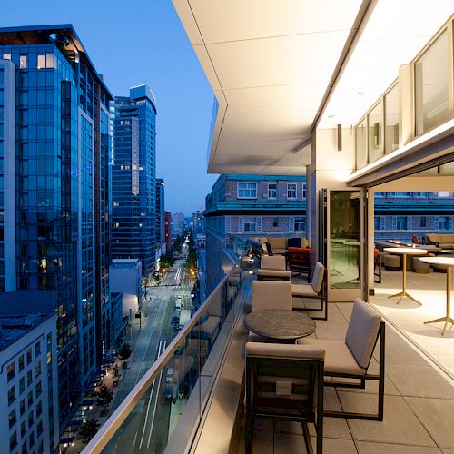 A modern balcony overlooks an urban street, lined with tall buildings, at dusk. The space features tables and chairs, with city lights visible.