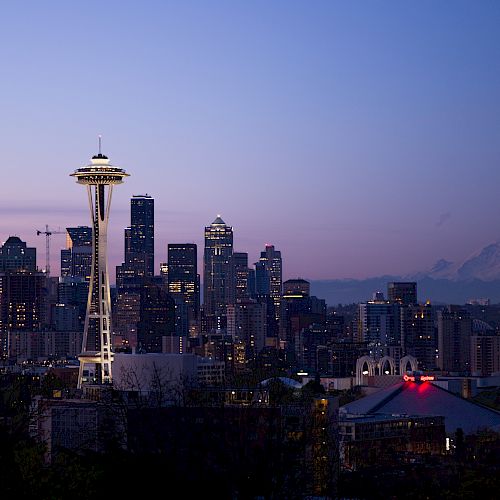A city skyline at dusk with the Space Needle and Mount Rainier visible in the background under a purple and pink sky.