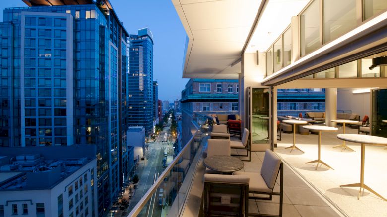 A modern rooftop terrace overlooking a cityscape with tall buildings at dusk, featuring tables and lounge chairs along a glass railing.
