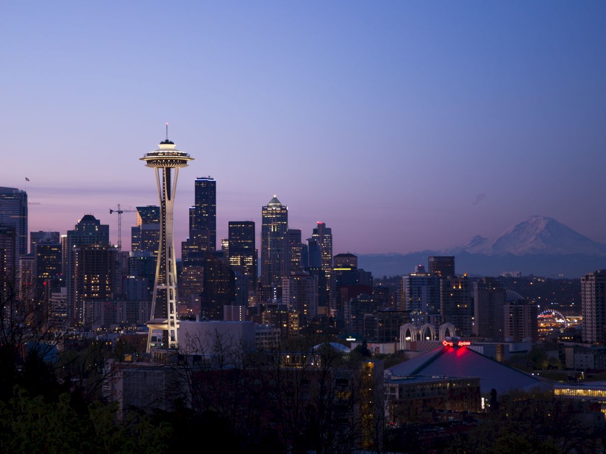 Seattle skyline at dusk with the Space Needle and Mount Rainier in the background, under a clear blue and purple sky.