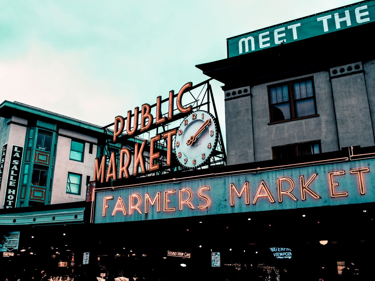 The image shows a neon sign for a "PUBLIC MARKET" and "FARMERS MARKET" with an analog clock, set against urban buildings.