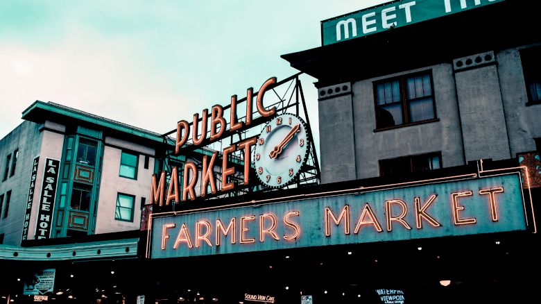 The image shows a neon sign for a "PUBLIC MARKET" and "FARMERS MARKET" with an analog clock, set against urban buildings.