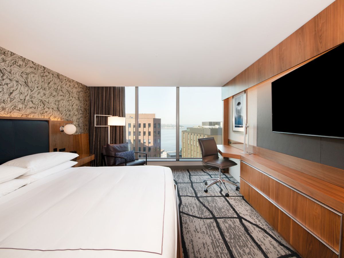 A modern hotel room with a large bed, TV, desk, chair, and a view of buildings and water through a wide window.